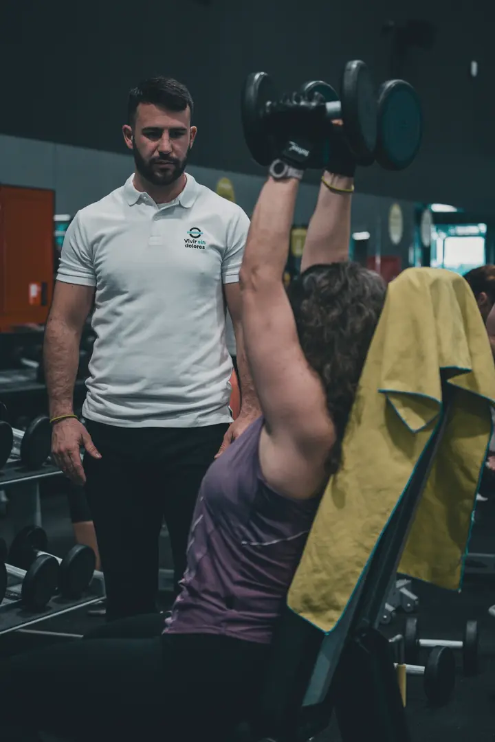 A personal trainer standing by as a client presses dumbbells overhead