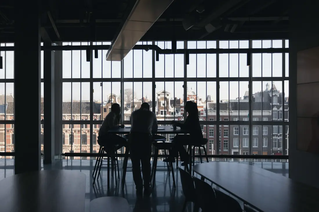 People meeting at a table beside tall windows overlooking the city